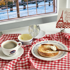 Breakfast scene with coffee, bagel, and cream cheese on a checkered tablecloth in front of a window view.